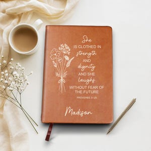 May include: A brown leather journal with the text "She is clothed in strength and dignity and she laughs without fear of the future" and the name "Madison". A floral design is on the left side. A pen and a cup of coffee are also in the image.