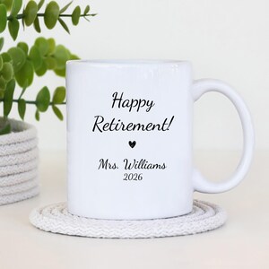 May include: White ceramic mug with the text "Happy Retirement!" in black script, a black heart, and "Mrs. Williams 2026". The mug is on a white knitted coaster, with a green plant in the background.