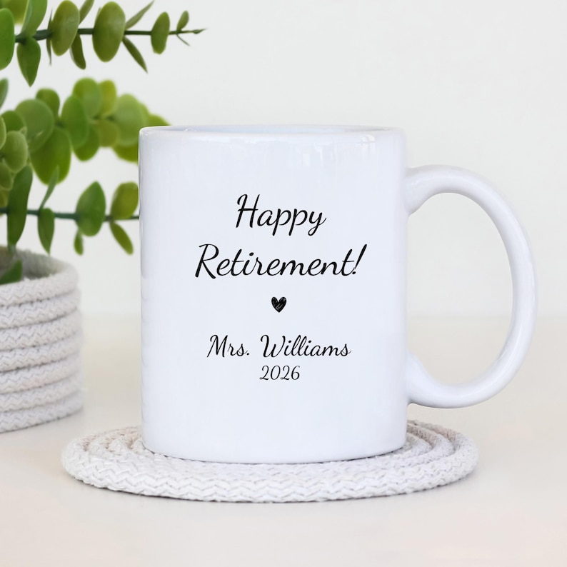 May include: White ceramic mug with the text "Happy Retirement!" in black script, a black heart, and "Mrs. Williams 2026". The mug is on a white knitted coaster, with a green plant in the background.