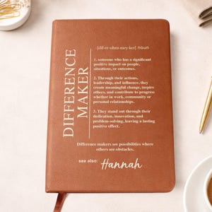 May include: A brown leather-bound journal with the words "DIFFERENCE MAKER" vertically down the left side. The journal includes a definition, and the name "Hannah" at the bottom. A bookmark is visible.
