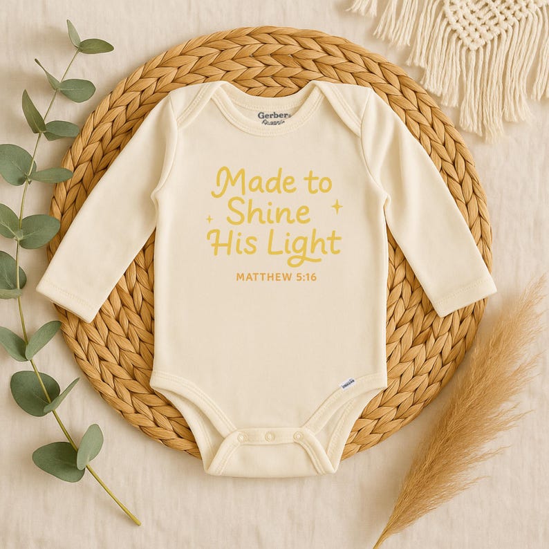 May include: Cream-colored baby onesie with long sleeves. The text "Made to Shine His Light" and "Matthew 5:16" are printed in yellow. The onesie is displayed on a woven, circular placemat with decorative elements.