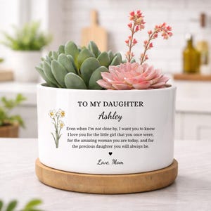 May include: A white ceramic planter with a succulent arrangement. The planter has text that reads "TO MY DAUGHTER Ashley" and a heartfelt message. The arrangement includes green and pink succulents, and sits on a wooden base.