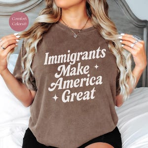 May include: A brown t-shirt with the text "Immigrants Make America Great" in white, with star accents. The shirt is a Comfort Colors brand.