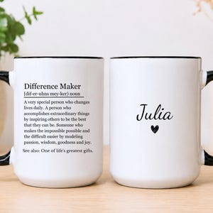 May include: Two white ceramic mugs with black handles and rims. One mug features the black printed text 'Difference Maker' followed by a dictionary-style definition. The other mug displays the name 'Julia' in black script above a small black heart.