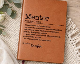 May include: A tan leather-bound notebook featuring the printed definition of the word Mentor, including the phonetic spelling, a list of descriptive phrases, and the text See also: Krsitin, with a dark maroon ribbon bookmark extending from the bottom.