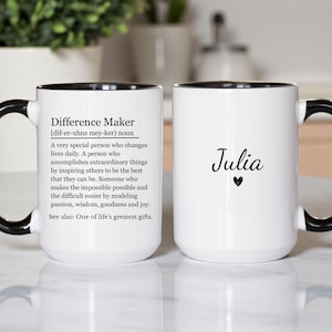 May include: Two white ceramic mugs with black handles and rims. One mug features the text "Difference Maker" with a definition. The other mug has the name "Julia" and a heart symbol.