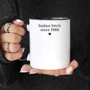 May include: White ceramic mug with a black interior and handle. The mug features the text "badass bitch since 1986" in black, with a small heart symbol below. The mug is held by a person wearing rings.