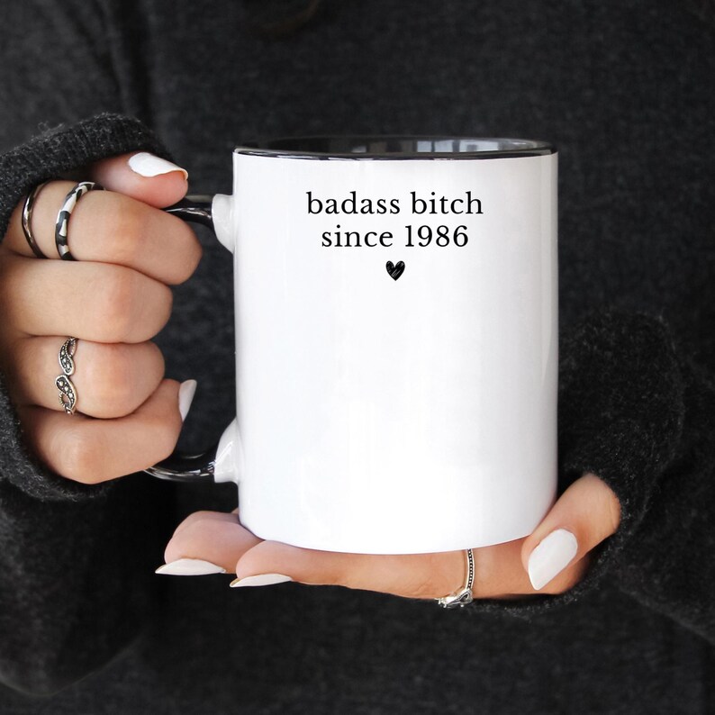 May include: White ceramic mug with a black interior and handle. The mug features the text "badass bitch since 1986" in black, with a small heart symbol below. The mug is held by a person wearing rings.