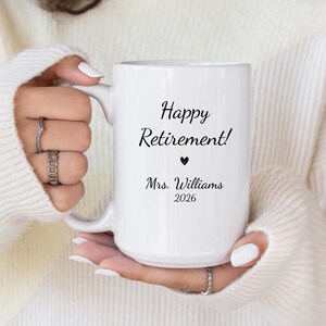 May include: White ceramic mug with the text "Happy Retirement!" in black script, a black heart, and the name "Mrs. Williams 2026". The mug is held by a person wearing a white sweater.