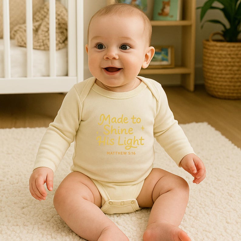 May include: A smiling baby wearing a long-sleeved, light yellow onesie. The onesie has the text "Made to Shine His Light" in gold lettering, with "Matthew 5:16" below. The baby is sitting on a cream-colored rug.