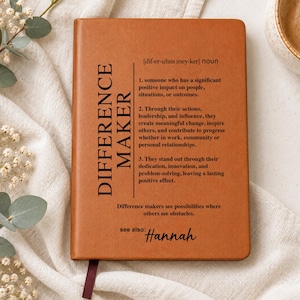 May include: A tan notebook with a smooth cover featuring the black printed definition of the noun Difference Maker, including numbered descriptions of the term, the phrase Difference makers see possibilities where others see obstacles, and see also: Hannah.