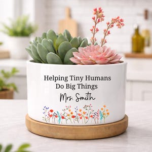 May include: A white ceramic planter with the text "Helping Tiny Humans Do Big Things Mrs. Smith" and a floral design. The planter holds a succulent and a pink flower, sitting on a wooden saucer.