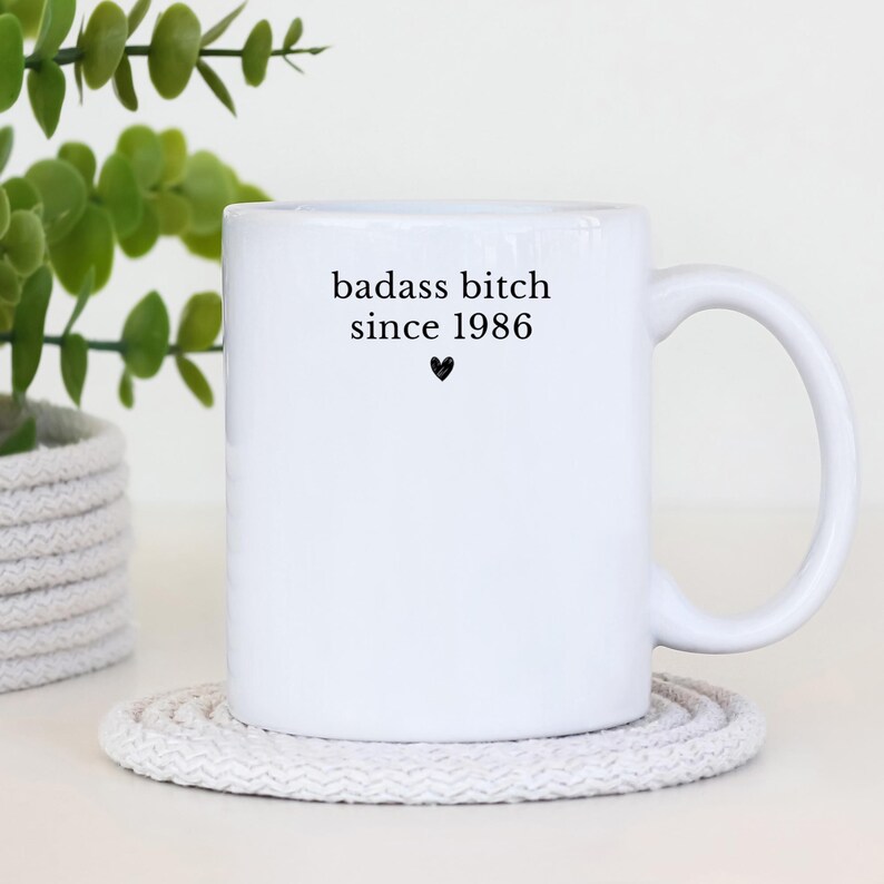 May include: A white ceramic mug with a black handle and the text "But First, Coffee" in a black, handwritten-style font. The mug is on a white surface.