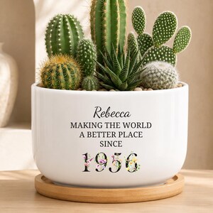 May include: A white ceramic planter containing a variety of green cacti and succulents, resting on a circular wooden tray. The planter features black text that reads Rebecca MAKING THE WORLD A BETTER PLACE SINCE 1956, with floral accents on the numbers.