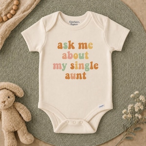 May include: A cream-colored short-sleeve baby bodysuit featuring the text "ask me about my single aunt" printed in a retro-style font with alternating colors of orange, pink, green, and yellow. The garment has a round neckline and snap closures.