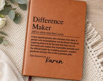 May include: A tan leather-bound notebook featuring black printed text that defines the term Difference Maker, followed by the name Karen. The notebook includes a thin, dark red ribbon bookmark extending from the bottom edge of the cover.