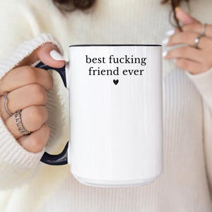 May include: White ceramic mug with a black interior and handle. The mug features the text "best fucking friend ever" in black, with a small black heart below the text. The mug is held by a person.