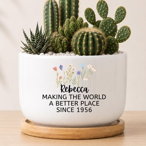 May include: A white ceramic planter with a bamboo saucer containing various green cacti and succulents. The planter features a colorful wildflower graphic above the black text: Rebecca, MAKING THE WORLD A BETTER PLACE SINCE 1956.