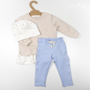 Könnte beinhalten: Ein Baby-Outfit-Set mit einem beigen Sweatshirt, hellblauen Jogginghosen und einer weißen Mütze mit goldenen Herzen. Das Sweatshirt hat einen weißen Rüschenbesatz und die Jogginghose hat einen Kordelzug.