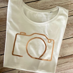 Könnte beinhalten: Weißes T-Shirt mit einer goldenen Umrandung einer Kamera.