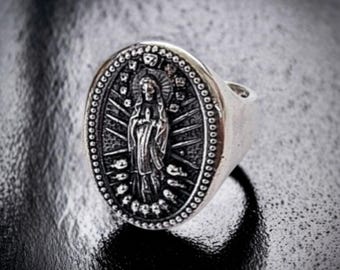 May include: A silver-toned metal ring featuring an oval face with a raised, detailed relief of a figure in robes with hands clasped in prayer, surrounded by a sunburst pattern and a beaded border, set against a dark, textured background.
