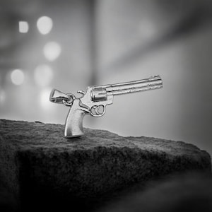 May include: A silver-coloured revolver is positioned on a dark, textured surface. The image is a close-up, highlighting the gun's detailed design and metallic finish. The background is blurred, drawing focus to the firearm's shape.