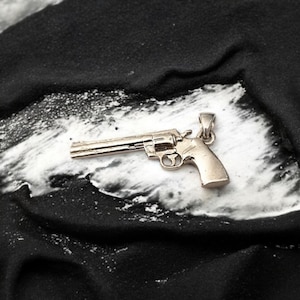 May include: A silver-colored revolver pendant. The detailed charm is a miniature replica of a classic handgun, featuring a cylinder, barrel, and handle. The pendant is displayed on a black and white textured surface.