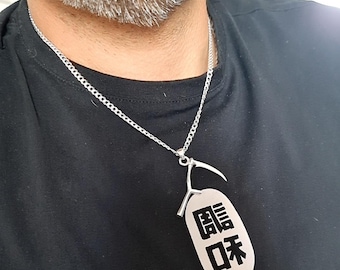 May include: A silver-toned metal necklace featuring a thin chain and a pendant shaped like a scythe attached to an oval-shaped tag. The tag displays two large black Japanese kanji characters printed vertically on its smooth, light-colored surface.