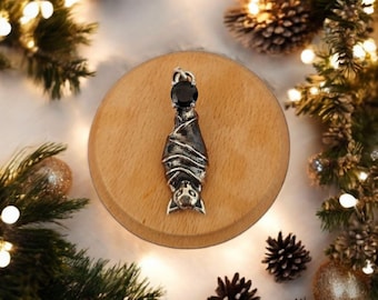 May include: A silver-toned metal pendant shaped like a bat hanging upside down, featuring a round, faceted black gemstone set at the top. The bat figure has detailed wings and facial features, with a small loop at the top for a chain.