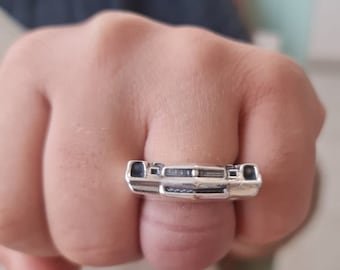 May include: A silver ring featuring a detailed, three-dimensional front view of a classic muscle car, including the grille, headlights, and bumper design. The metallic finish has dark recessed areas to highlight the intricate automotive features.