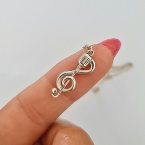 May include: A silver-colored pendant necklace featuring a treble clef intertwined with a vintage microphone design. The pendant is small and detailed, with a delicate chain. The necklace is likely made of sterling silver or a similar metal.