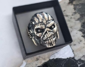 May include: A silver-toned metal ring featuring a detailed, sculpted skull design with deep-set eyes, prominent teeth, and textured hair-like grooves. The ring is presented inside a small, square black box with a smooth, matte finish.