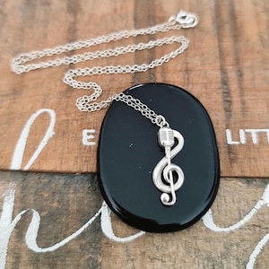 May include: A silver necklace featuring a treble clef and microphone pendant. The pendant is attached to a silver chain and rests on a black oval stone. The background is a wooden surface with white lettering.