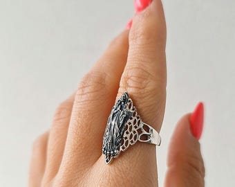 May include: A silver ring featuring a tall, pointed centerpiece with a dark, oxidized relief of a figure in flowing robes. The band is constructed from an intricate, openwork filigree pattern that extends from the central design around the finger.