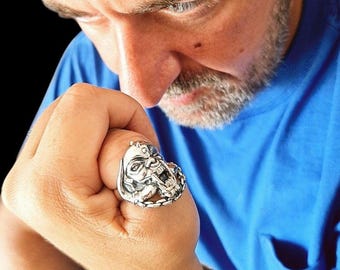 May include: A silver ring featuring a detailed, sculpted creature head with prominent teeth and textured facial features. The band of the ring has a braided or chain-link design that wraps around the finger, showcasing a metallic finish.