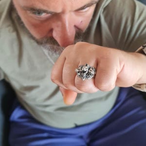 Könnte beinhalten: Ein silberfarbener Metallring mit einem detaillierten Design einer dreiköpfigen Kreatur. Der Ring hat eine polierte Oberfläche mit dunklen, vertieften Bereichen, die die komplexen skulpturalen Merkmale der nebeneinander liegenden Köpfe betonen.