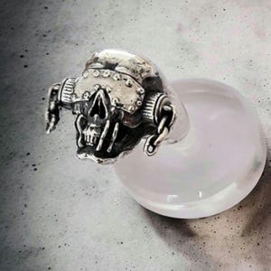 May include: A silver skull ring with intricate detailing. The skull features a helmet design with rivets and chain accents. The ring is mounted on a clear, rounded base, possibly for display or photography purposes.