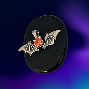 May include: A silver bat pendant with an orange gemstone centre is displayed on a black oval. The bat's wings are outstretched, and the gemstone is round. The background is a gradient of blue and purple.