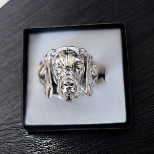 May include: A silver-toned ring featuring a detailed, three-dimensional sculpture of a dog head with long, drooping ears. The metal surface has a polished finish with darkened recessed areas to highlight the facial features of the canine design.