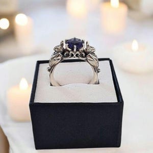 Könnte beinhalten: Ein silberner Ring mit einem großen, runden, dunkelvioletten Edelstein. Der Ring hat ein verziertes Design mit Blatt- und Blumendetails. Er wird in einer schwarzen Ringschatulle mit weißem Innenraum präsentiert, mit verschwommenen Kerzen im Hintergrund.
