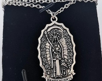May include: A silver-toned metal pendant necklace featuring an oval-shaped charm with a raised relief of Santa Muerte holding a scythe and scales. The border is decorated with roses, and the text SANTA MUERTE is engraved at the bottom.