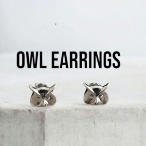 Op de afbeelding: Een paar zilverkleurige oorknopjes in de vorm van uilenkoppen met puntige oren en ronde ogen. De tekst OWL EARRINGS staat in het zwart boven de oorbellen gedrukt. De oorbellen hebben een metallic afwerking en een gedetailleerd ontwerp.