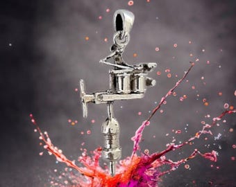 May include: A silver-colored pendant in the shape of a tattoo machine. The pendant has intricate details and is suspended from a small loop. The background is dark with colorful splashes of pink, orange, and red.