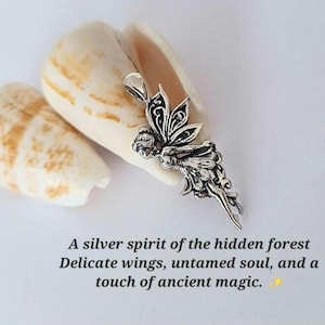 May include: A silver-toned metal pendant shaped like a winged figure with intricate filigree detailing on the wings and body. The pendant is positioned against a light-colored, textured shell. The text reads: A silver spirit of the hidden forest.