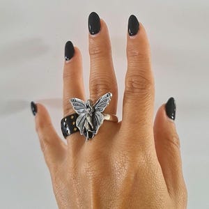 May include: A silver ring featuring a fairy with outstretched wings. The fairy ring is on a finger next to a black ring with gold-colored dots. The person's nails are painted black.