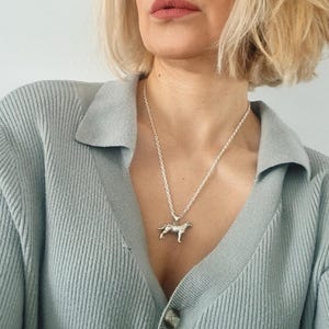 Könnte beinhalten: Halskette mit einem silberfarbenen Anhänger in Hundeform. Der Anhänger, in Form eines Hundes, hängt an einer silbernen Kette. Die Halskette wird über einer hellblauen Strickjacke getragen. Der Anhänger stellt einen Labrador oder eine ähnliche Rasse dar.