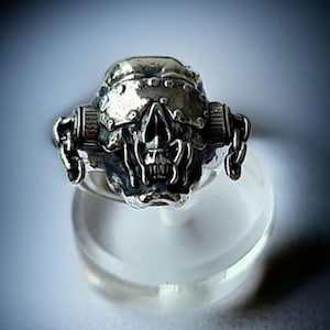 Può includere: Un anello a teschio d'argento con un design dettagliato. Il teschio ha un elmetto ed è affiancato da elementi cilindrici con catene. L'anello è esposto su un supporto trasparente e rotondo, mettendo in risalto l'intricata lavorazione e il tema gotico.