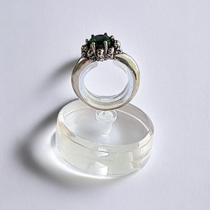 May include: A silver ring featuring a large, round, dark green gemstone. The ring is decorated with small skull motifs around the central stone. It's displayed on a clear, oval stand against a white backdrop.