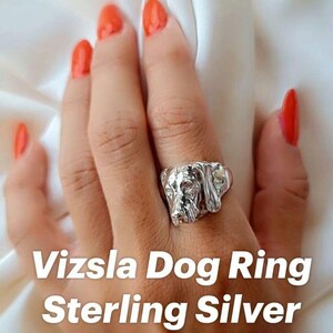 Puede incluir: Un anillo de plata de ley que presenta una escultura tridimensional detallada de una cabeza de perro Vizsla. El metal tiene un acabado pulido y reflectante que resalta las facciones del canino, incluyendo las orejas y el hocico.