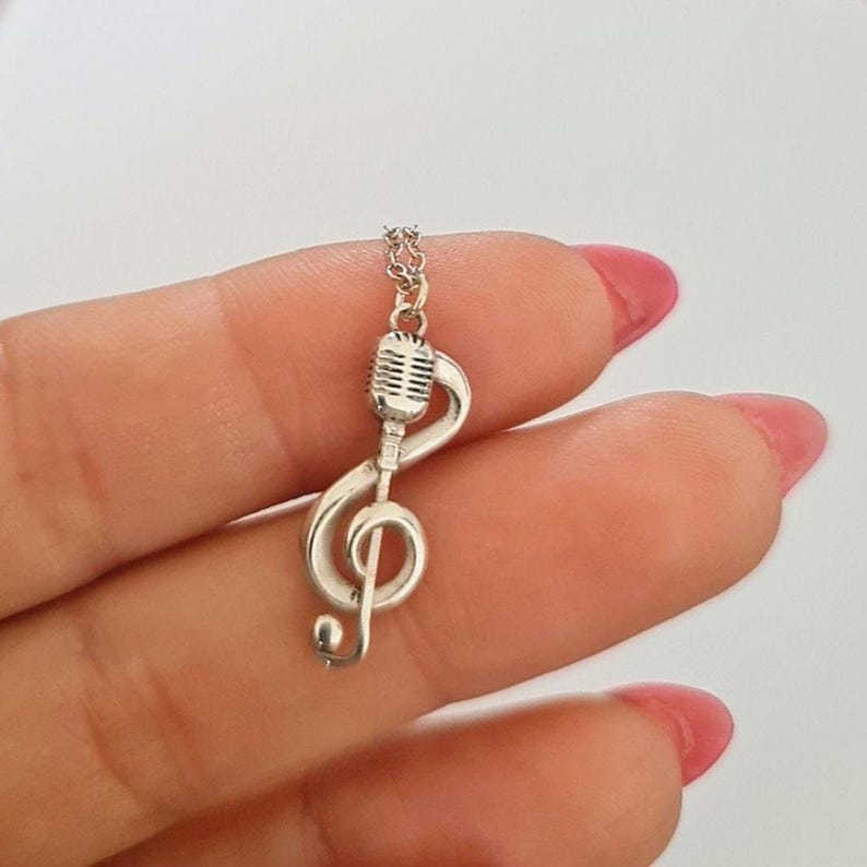 May include: A silver-colored necklace pendant featuring a treble clef combined with a vintage microphone design. The pendant hangs from a delicate chain. The jewelry is likely made of sterling silver or a similar metal. The design is suitable for music lovers.